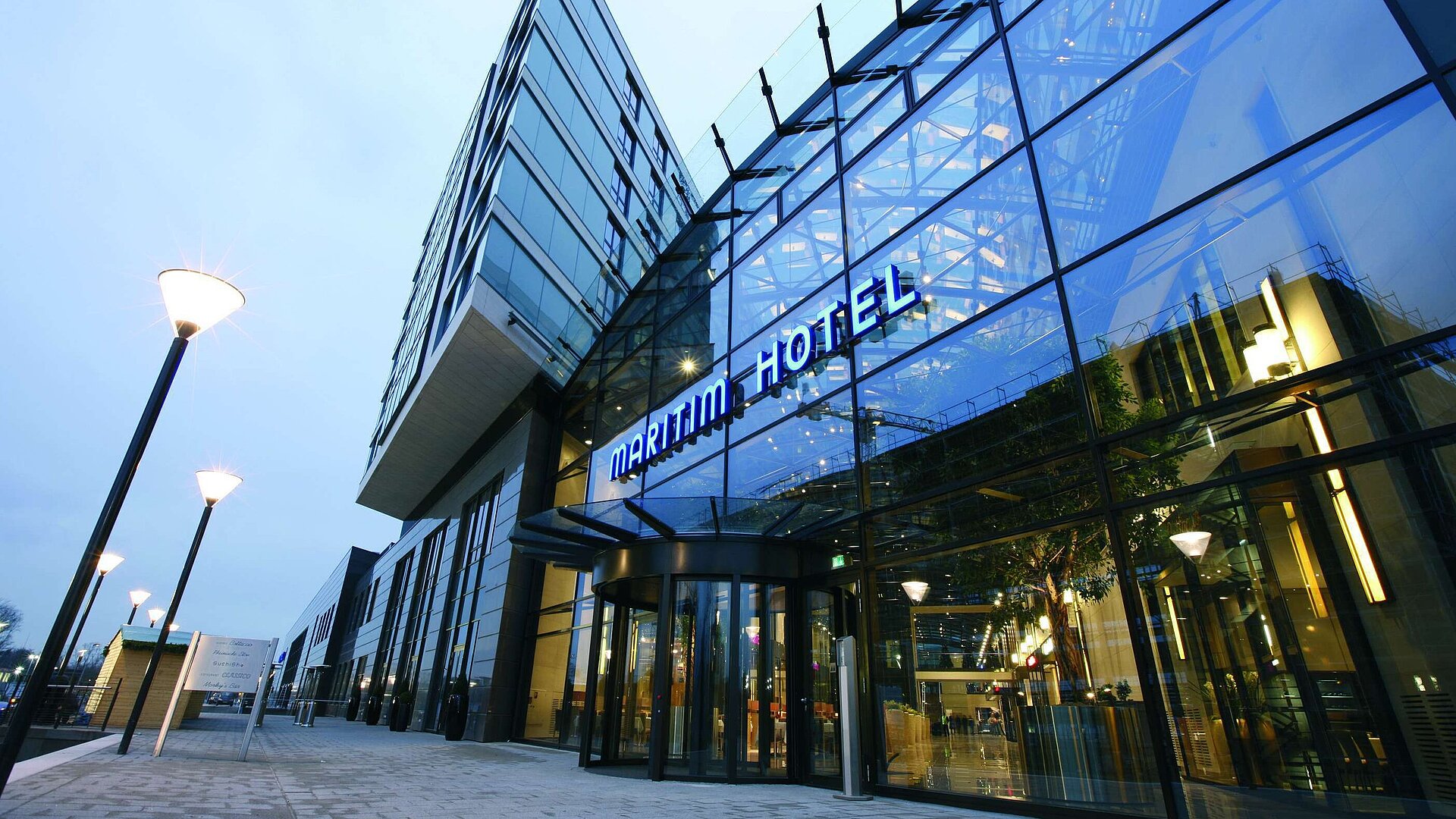 "Moderne Außenansicht des Maritim Hotel Düsseldorf mit gläserner Fassade und leuchtendem Hotellogo bei Dämmerung.