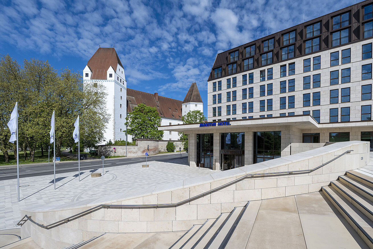 Das moderne Maritim Hotel Ingolstadt mit dem historischen Neuen Schloss im Hintergrund und einladendem Eingangsbereich.